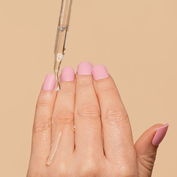 Hand with pink nail polish holding a dropper with clear liquid against a beige background